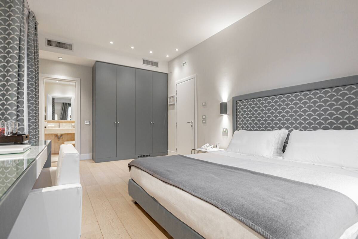 A modern bedroom with a large bed, grey wardrobe, and a glass dresser.