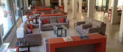 Een ruime zithoek met diverse zitplaatsen, waaronder banken en fauteuils, rondom een centrale salontafel. Er staan verschillende tafels en stoelen en in de achtergrond is een open haard te zien.