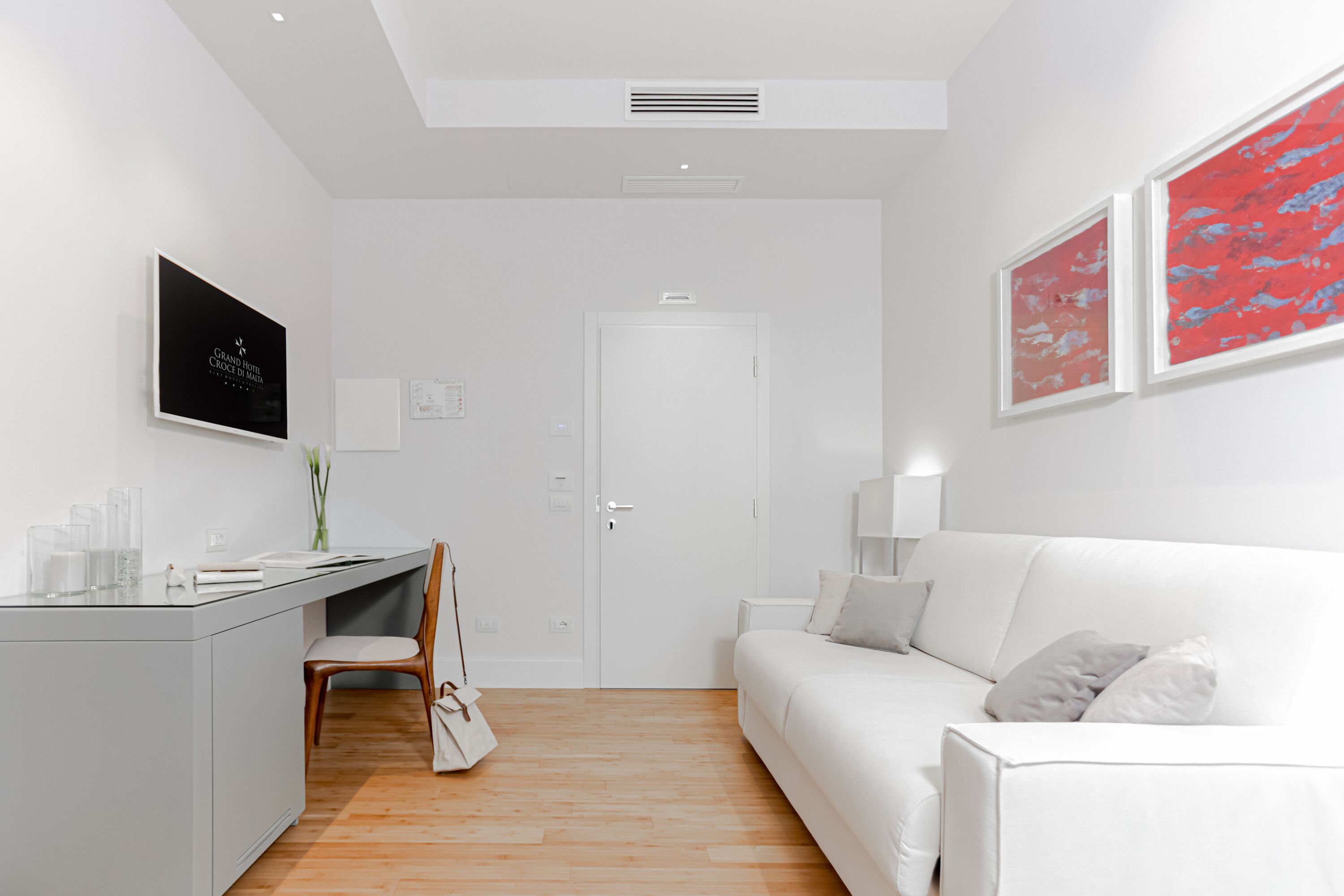 A modern, minimalist room with a white desk, chair, and sofa, a white door, and two framed abstract paintings.