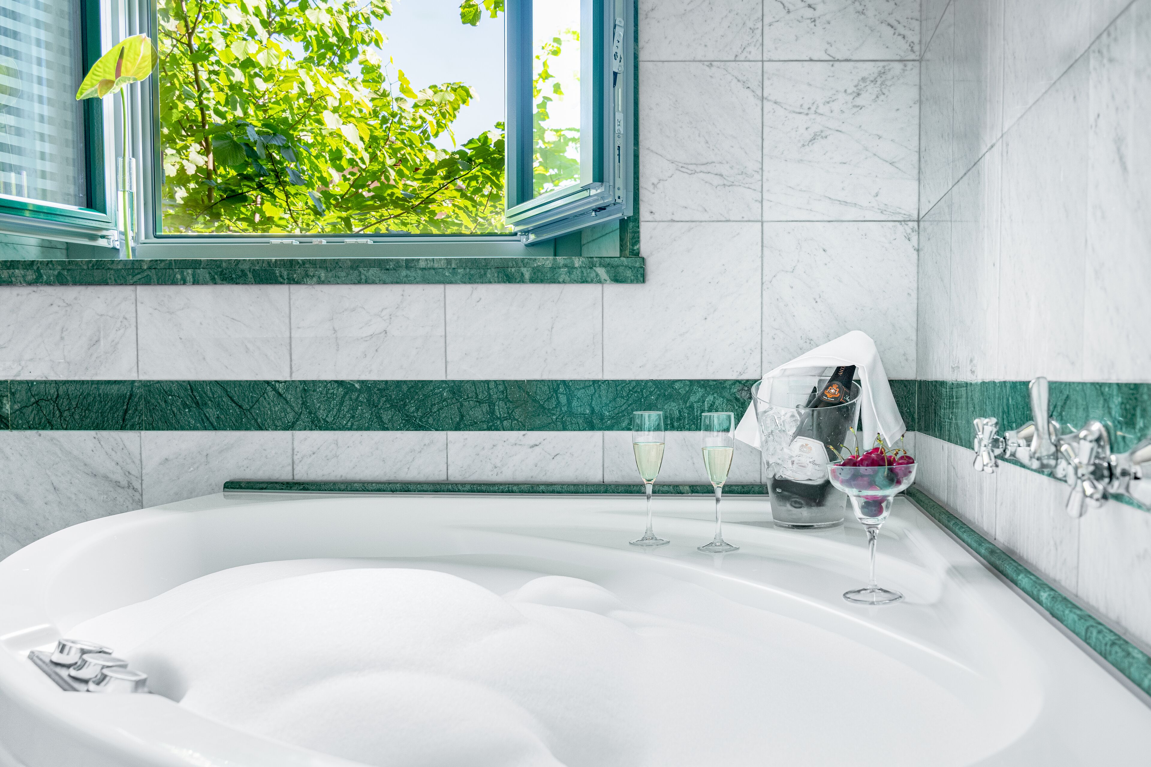 A modern bathroom with a large bathtub, a window with a view of green foliage, and a marble-tiled wall.