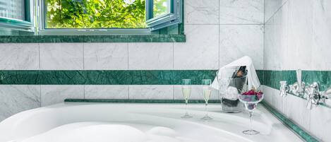 A modern bathroom with a large bathtub, a window with a view of green foliage, and a marble-tiled wall.