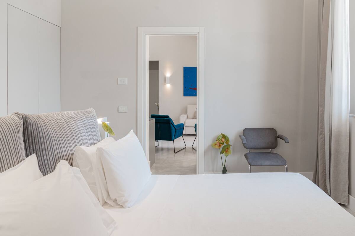 A hotel room with a bed, two chairs, a small table, and a view of the hallway.
