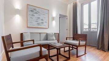 A modern living room with a sofa, two wooden chairs, and a coffee table. There is a framed artwork on the wall and a sliding glass door leading to a balcony.