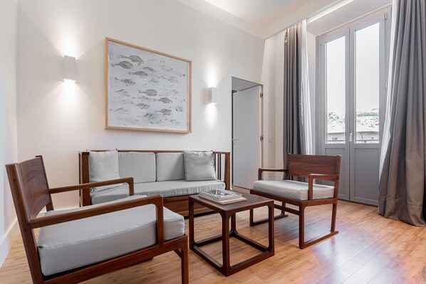 A modern living room with a sofa, two wooden chairs, and a coffee table. There is a framed artwork on the wall and a sliding glass door leading to a balcony. - Grand Hotel Croce di Malta Wellness & Golf (Montecatini Terme)