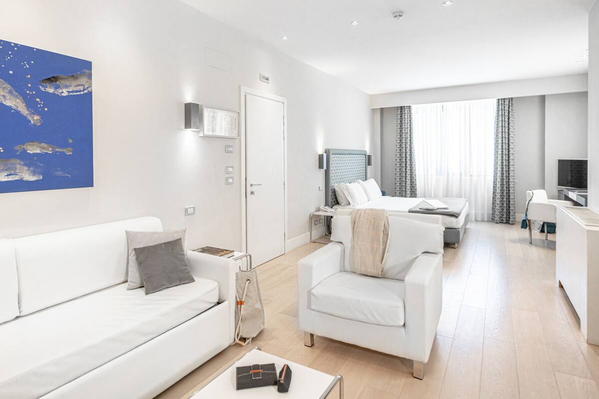 A modern hotel room with a white sofa, a smaller white armchair, a bed with a headboard, a desk, a television, and a large window with curtains.