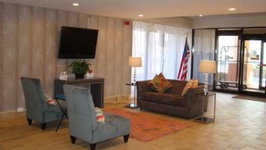 A hotel lobby with a grey sofa, two armchairs, a flat-screen TV, a potted plant, and an American flag. - Quality Inn West Springfield (West Springfield)