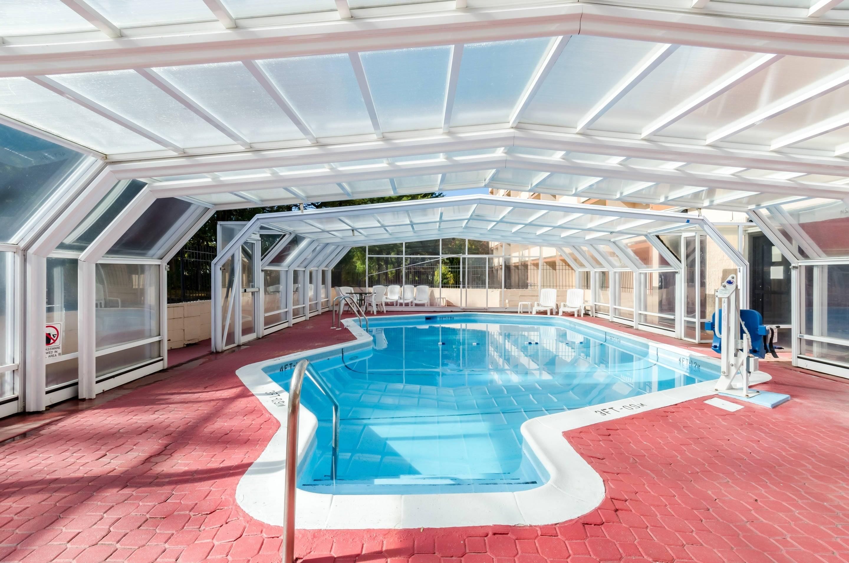 An indoor swimming pool with a glass ceiling, red flooring, and white poolside chairs.