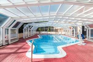 An indoor swimming pool with a glass ceiling, red flooring, and white poolside chairs.