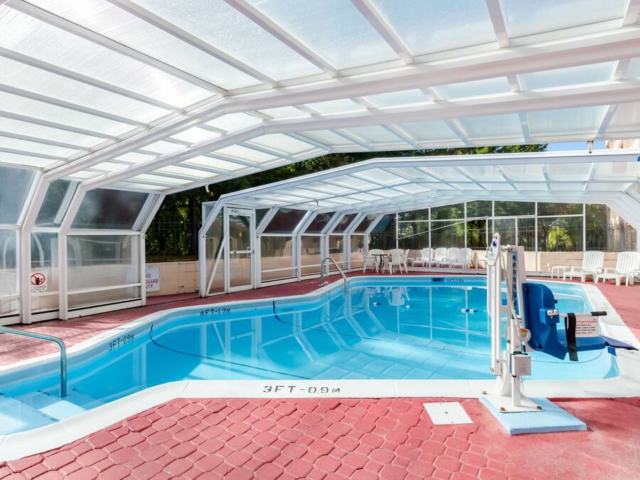 A covered swimming pool with glass walls, a diving board, and lounge chairs.