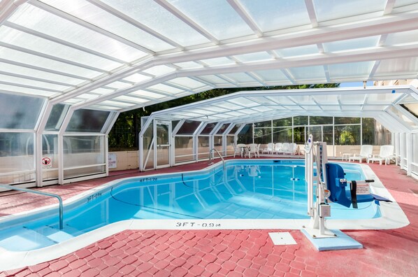 A covered swimming pool with glass walls, a diving board, and lounge chairs.