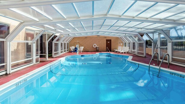 An indoor swimming pool with a glass ceiling, surrounded by white chairs and a wooden wall in the background.