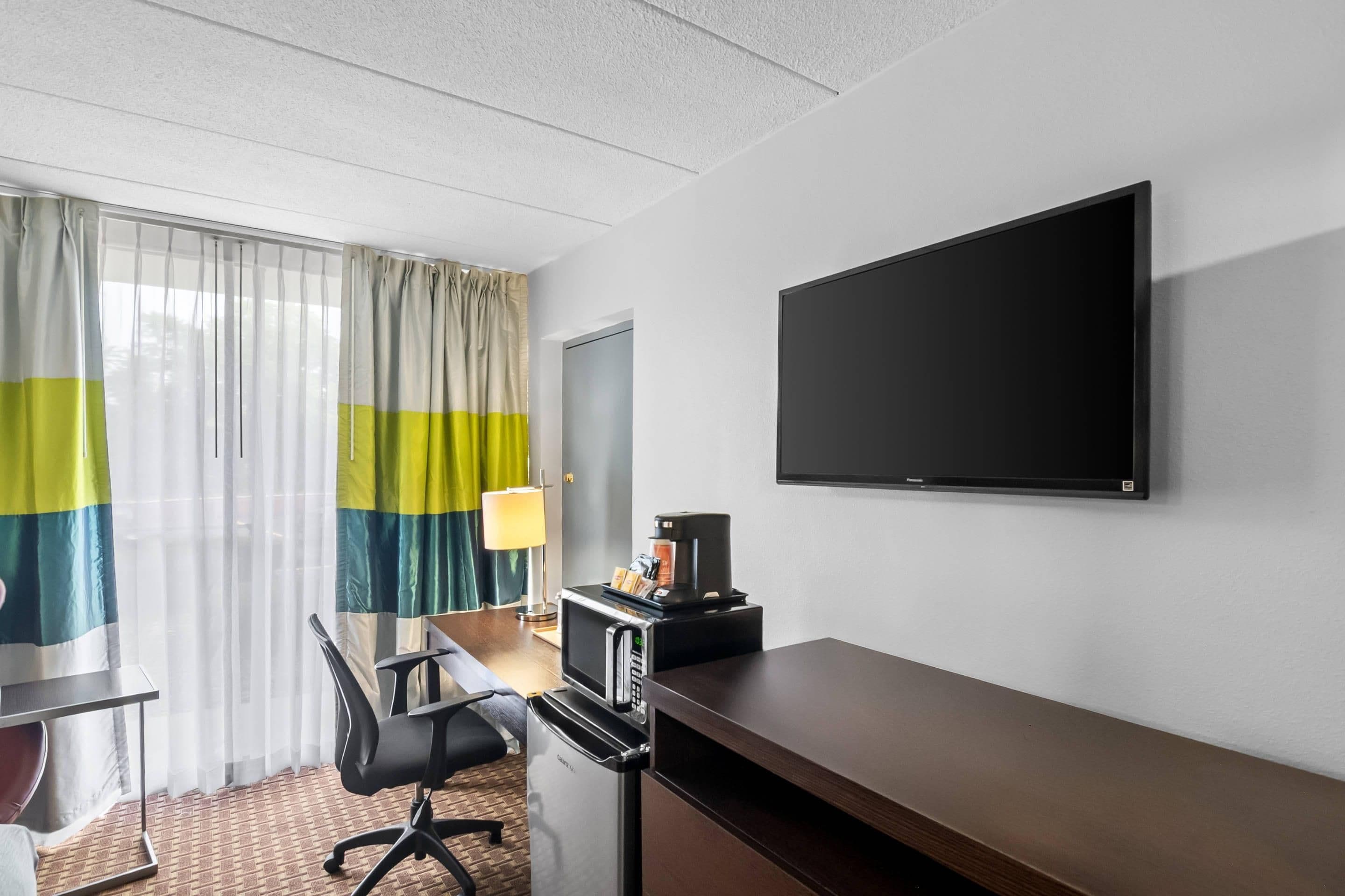 Quarto de hotel com cama, escrivaninha com cadeira, TV de tela plana, cafeteira e janela com cortinas.