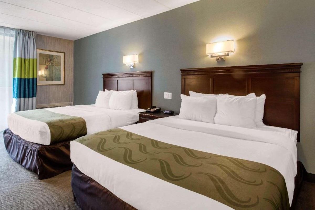 Habitación de hotel con dos camas, cada una con cabecero de madera, ropa de cama blanca y una manta estampada verde. Hay lámparas fijadas a la pared y un cuadro enmarcado.