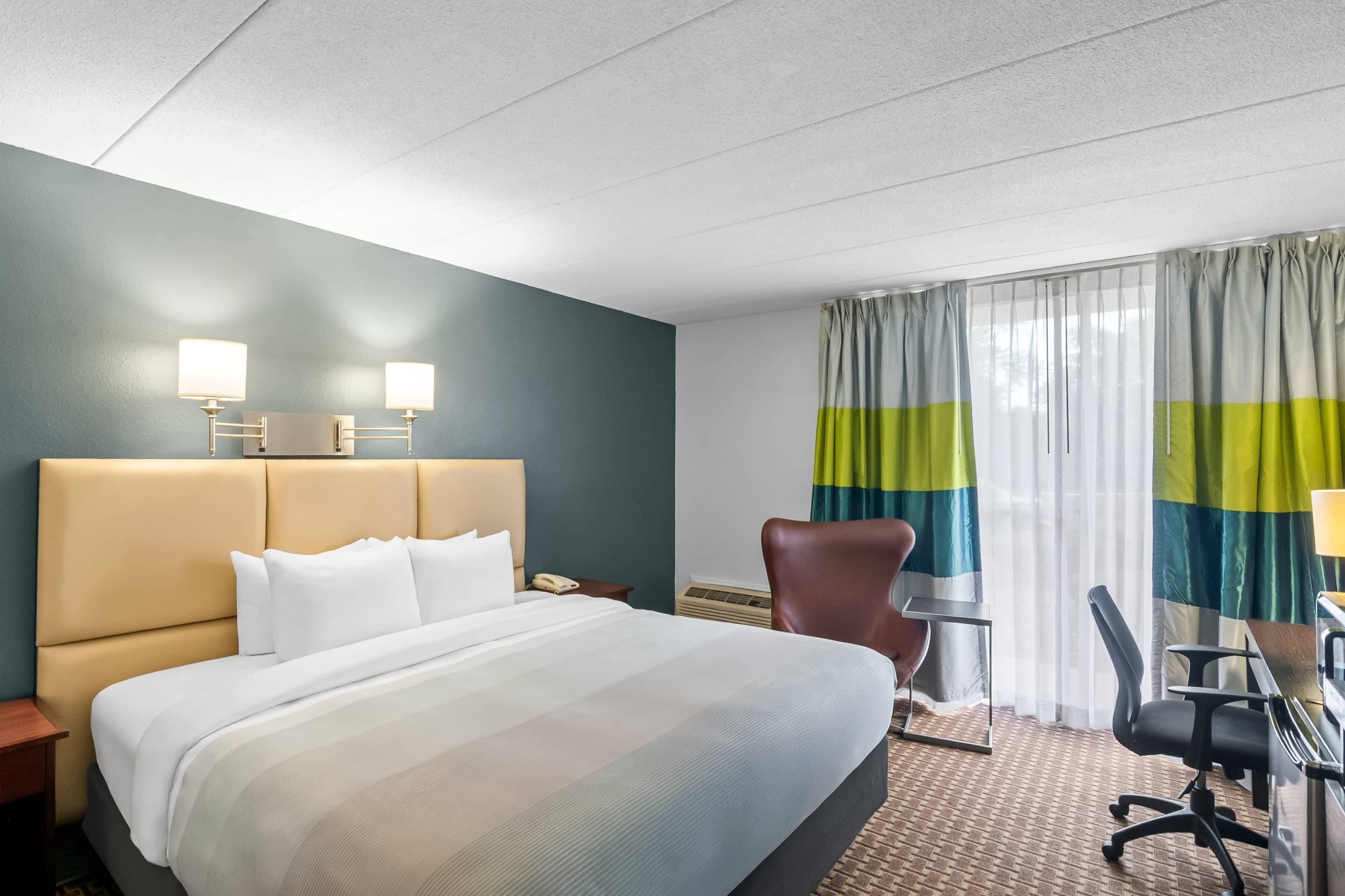 A hotel room with a large bed, a desk, a chair, a lamp, and a window with curtains.