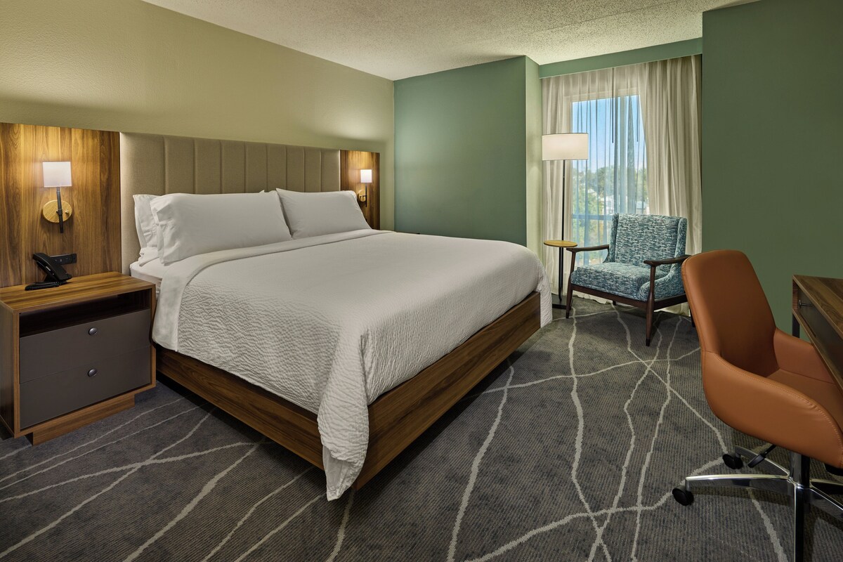 Quarto de hotel com uma cama grande, uma escrivaninha, uma cadeira e uma janela com cortinas.