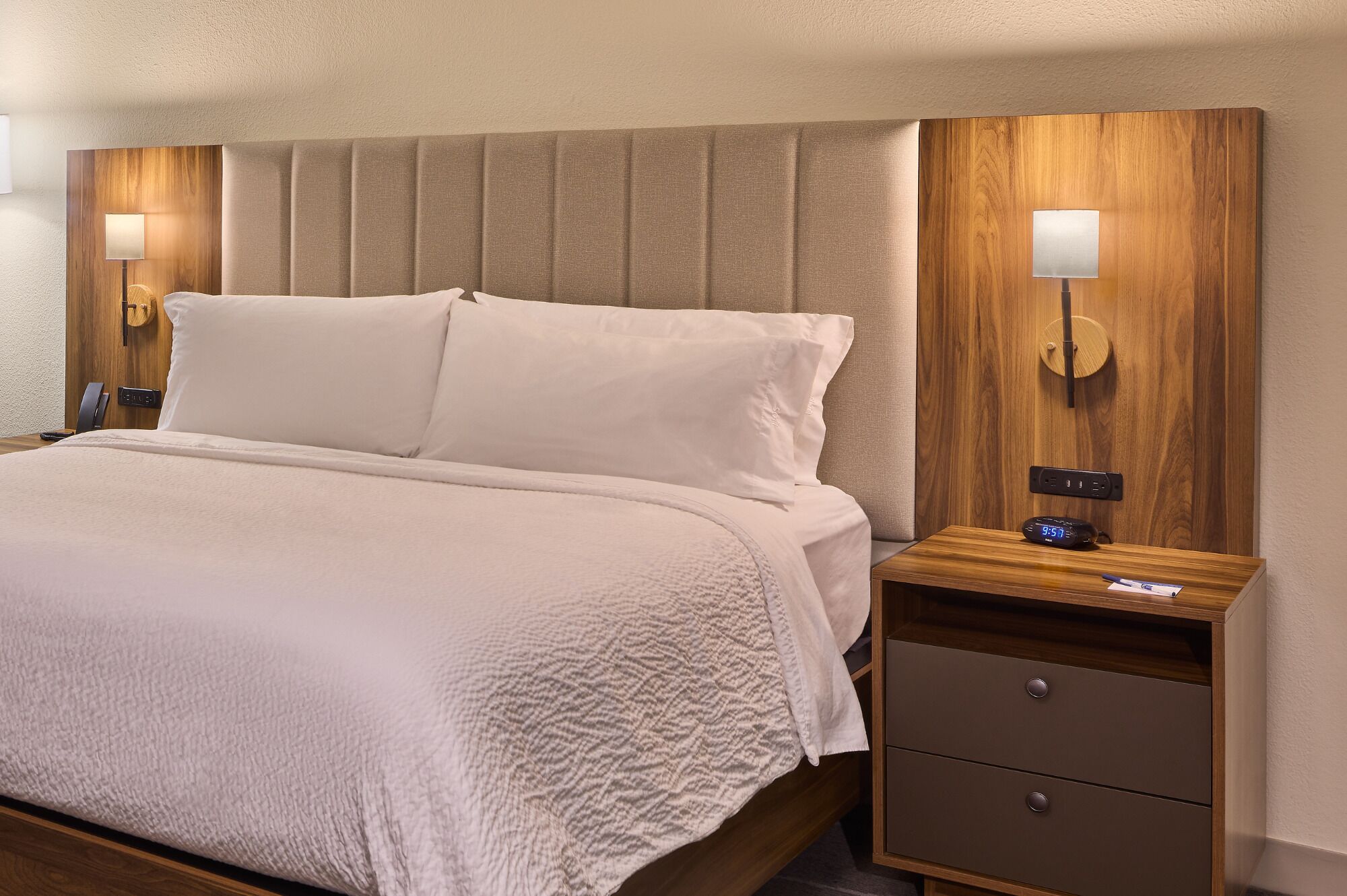 A neatly made bed with white linens, a wooden headboard, bedside lamps, a nightstand with a clock, and a drawer unit.
