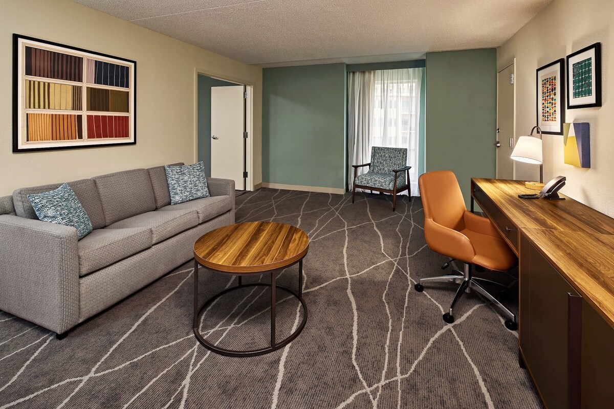 Quarto de hotel com sofá, mesa redonda de madeira, cadeira de escritório laranja, escrivaninha com abajur e tapete estampado.