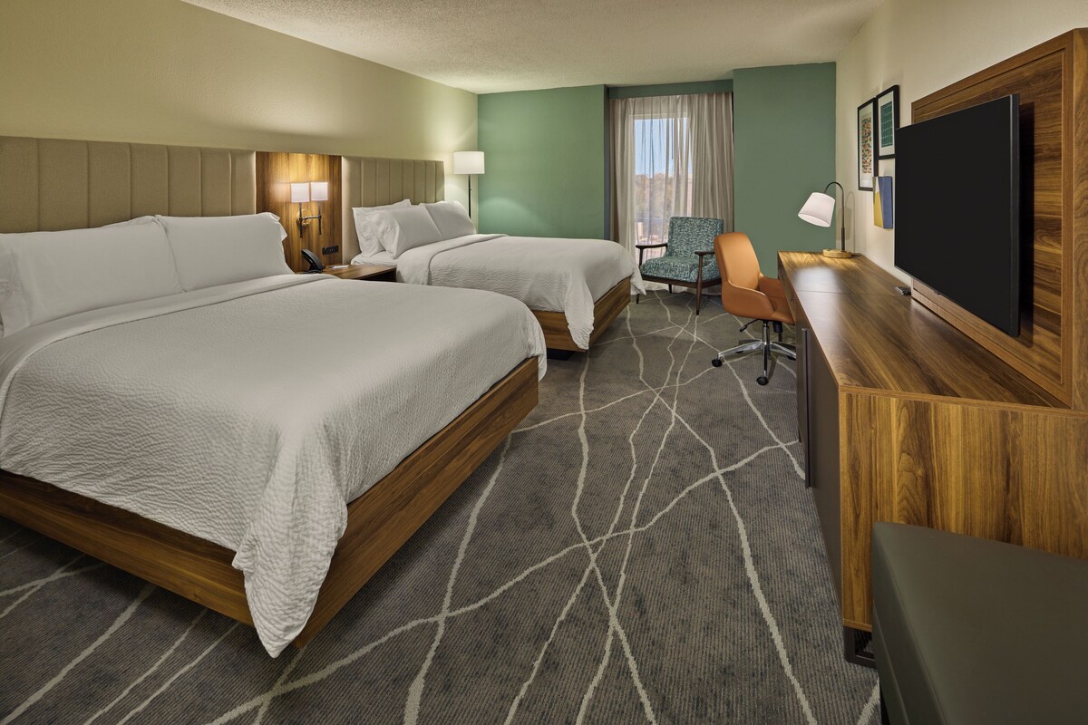 Quarto de hotel com duas camas, uma escrivaninha, uma cadeira, televisão e janela com cortinas.