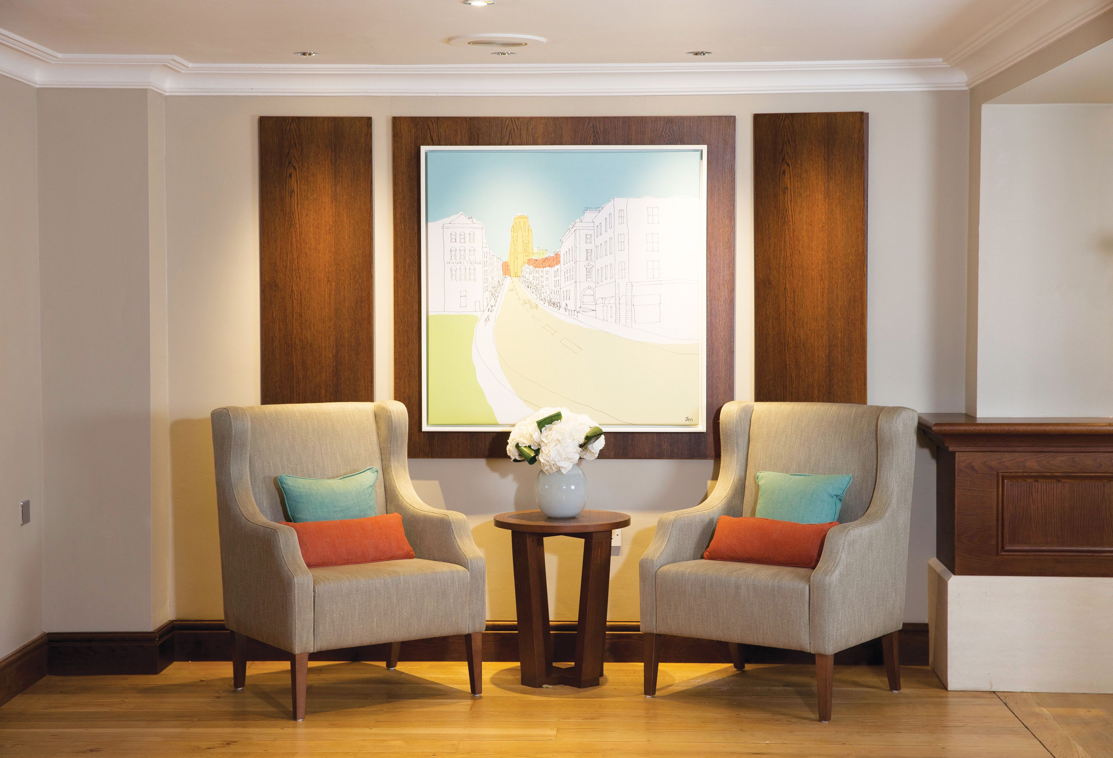 A room with two armchairs, a small table with a vase of flowers, and a large painting on the wall.