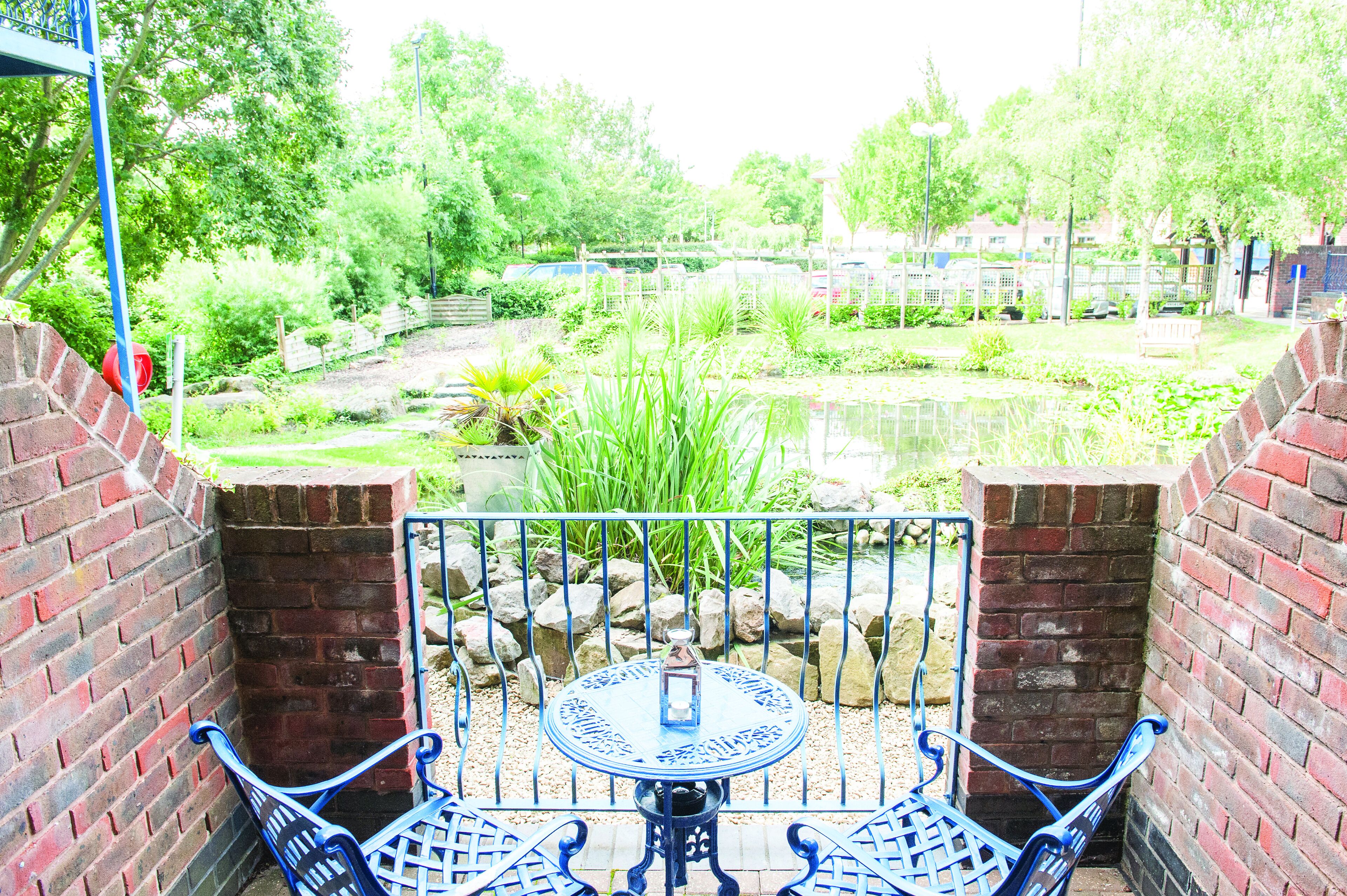 Un patio jardín con una mesa redonda y sillas de metal azules, rodeado de muros de ladrillo y un pequeño estanque.