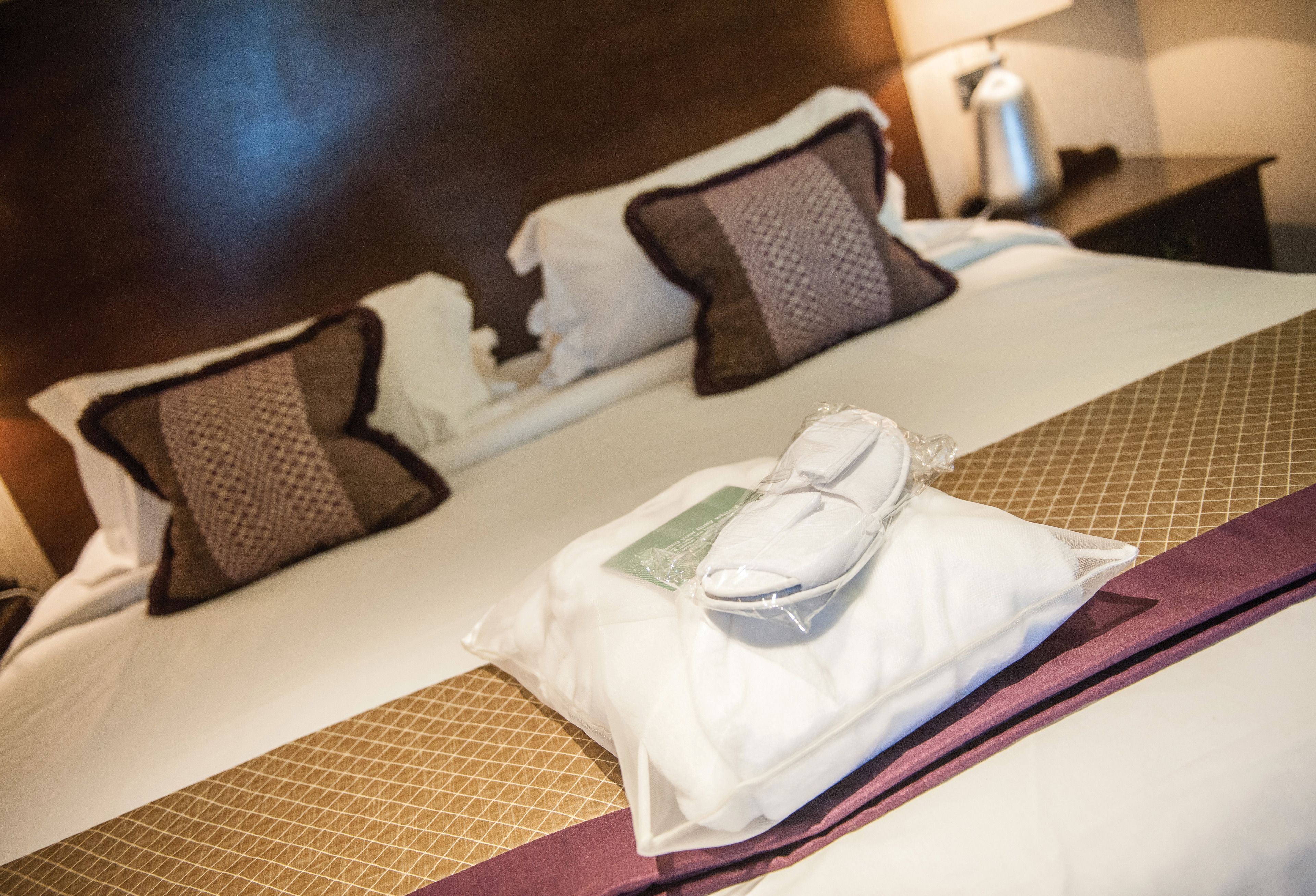 A neatly made bed with white linens, two brown pillows, and a pair of white slippers on a purple towel.