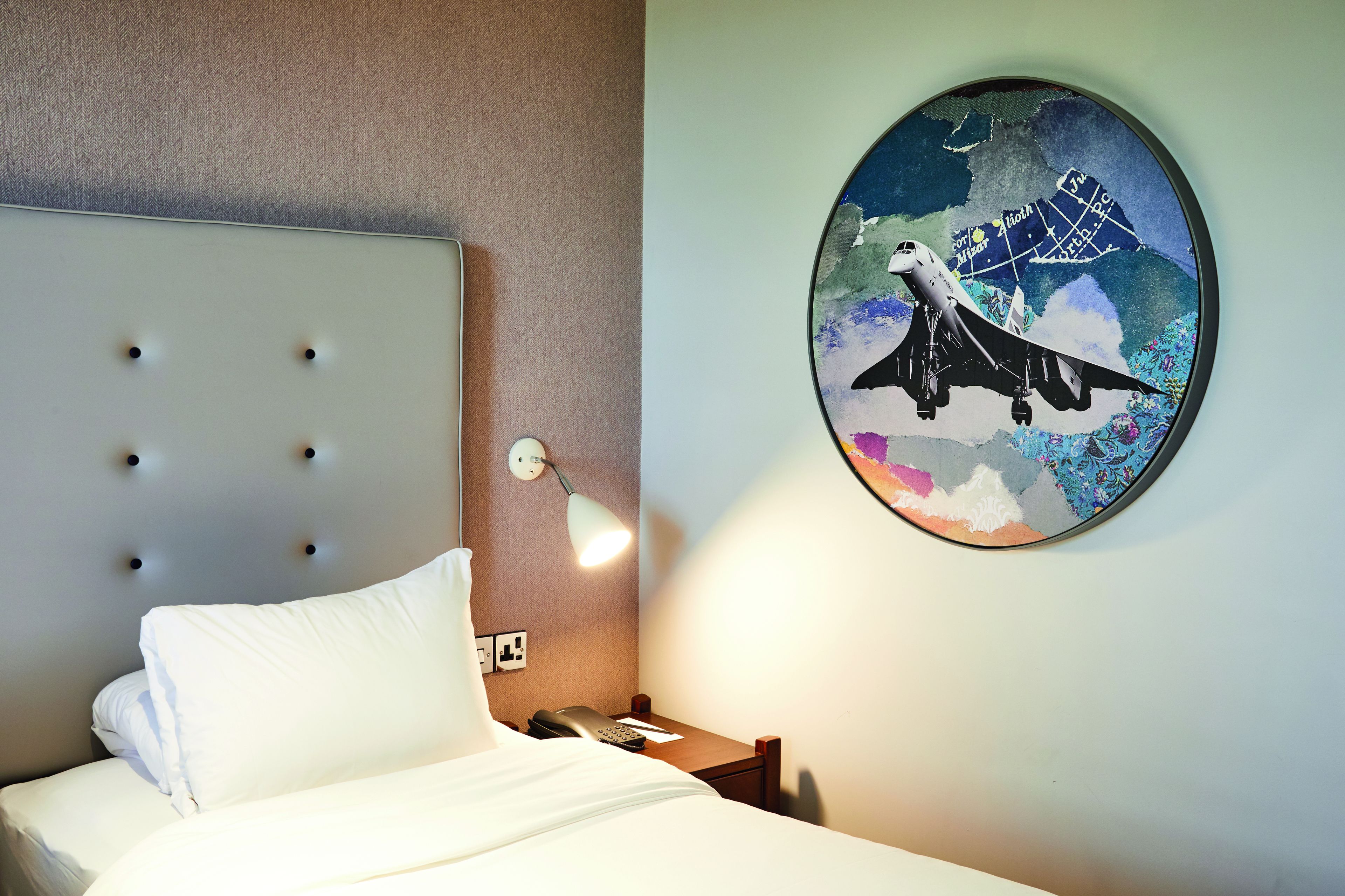 A hotel room with a bed, a nightstand, a lamp, and a wall-mounted artwork of an airplane.