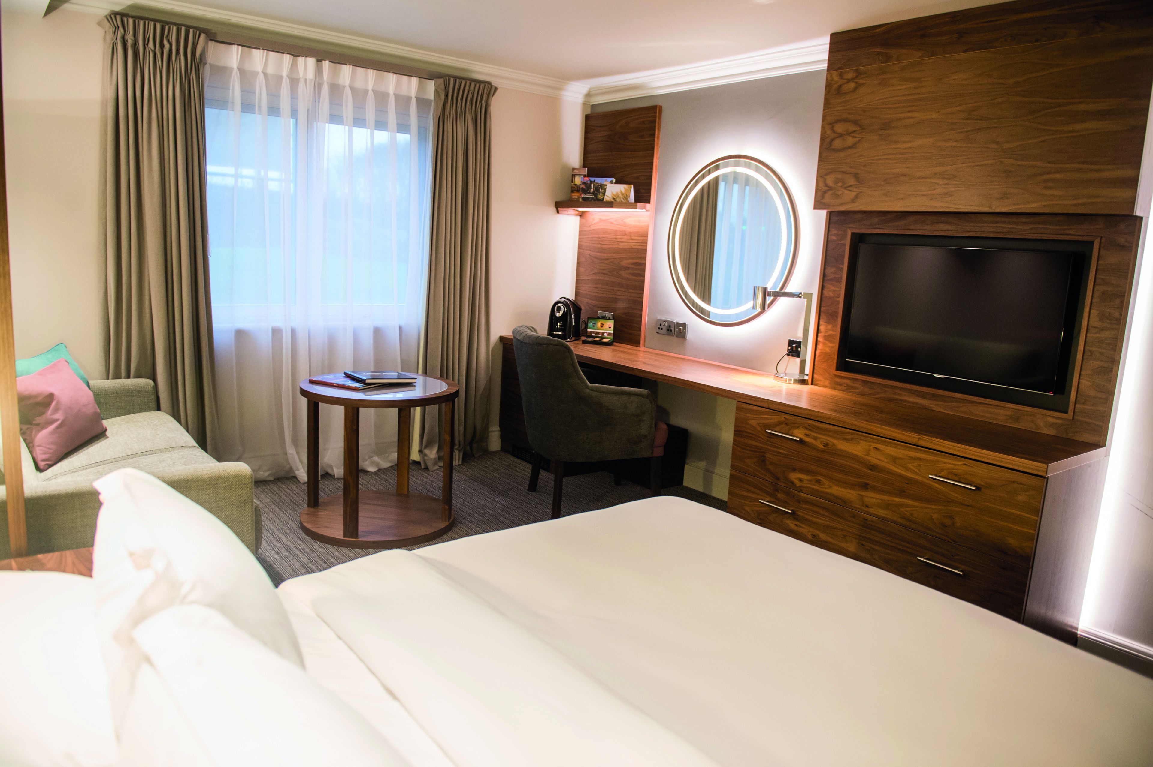 Une chambre d’hôtel comprenant un lit, un bureau avec une chaise, une télévision, un miroir et une fenêtre avec des rideaux.