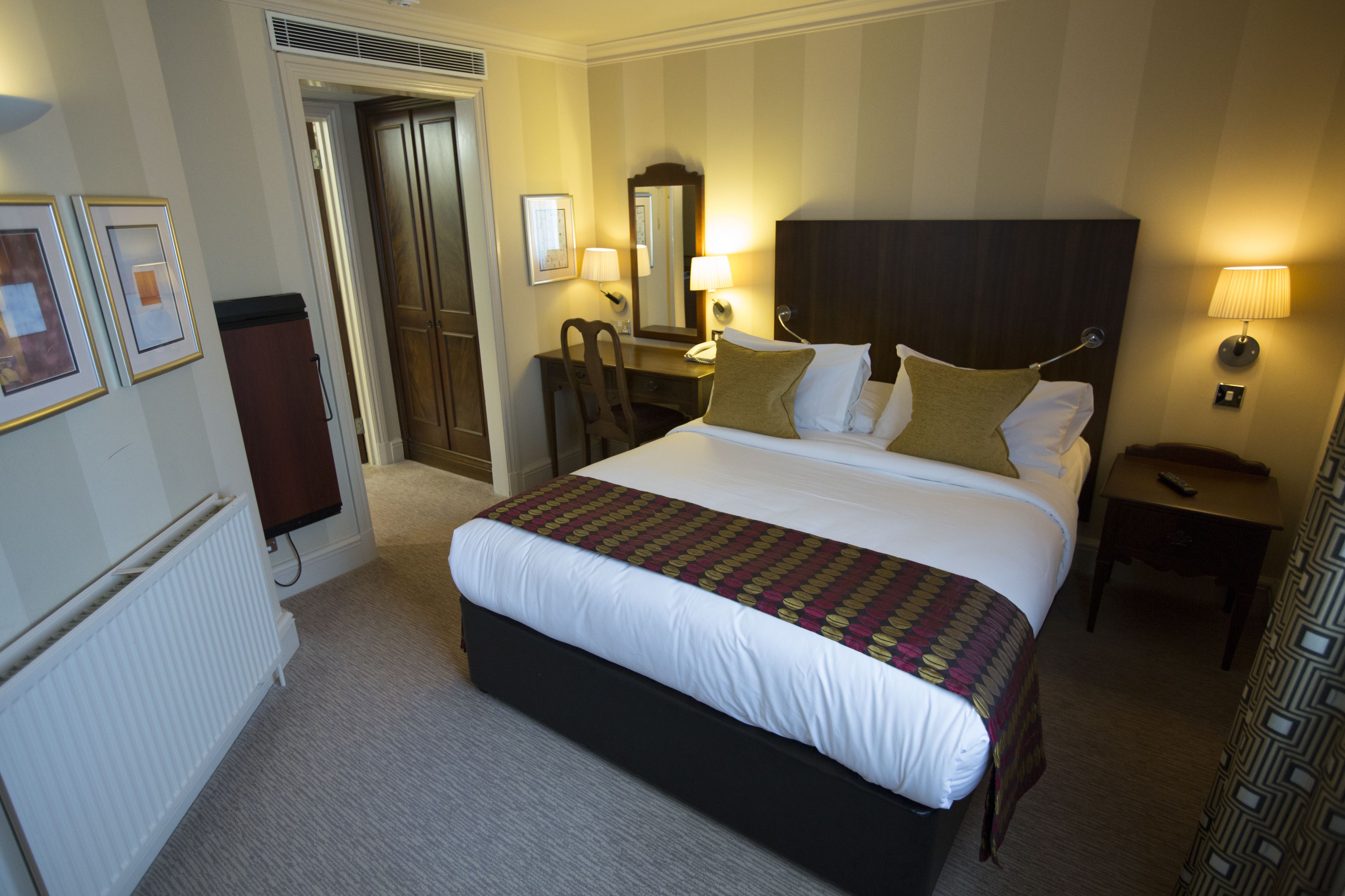 Una habitación de hotel con una cama grande, un escritorio con silla, un aparador, un espejo y una mesita de noche con lámpara.