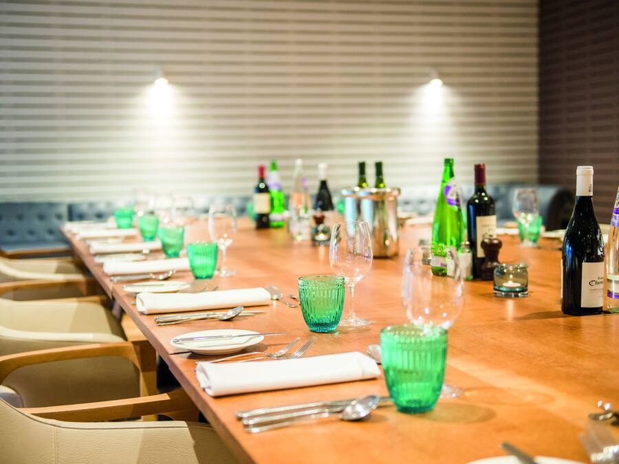 Una larga mesa de comedor dispuesta con cristalería verde, platos y cubiertos, con botellas de vino y copas sobre la mesa.