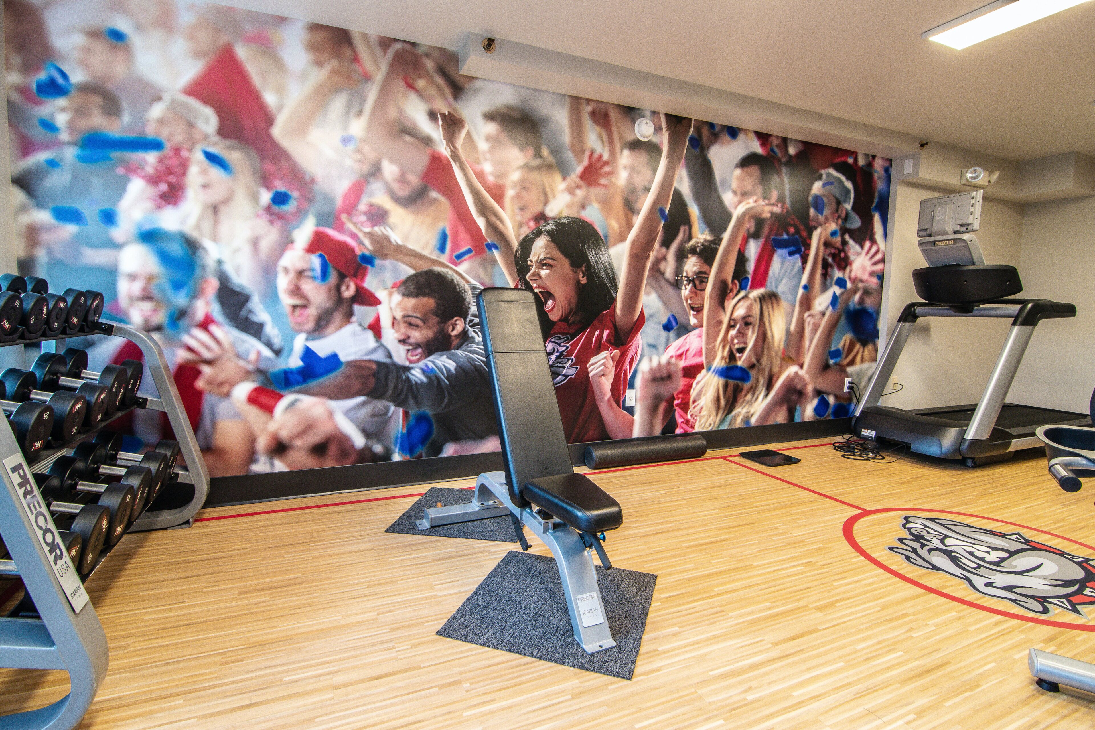 Un gimnasio con equipamiento de ejercicio, una pared con una multitud de personas y un logotipo en el suelo.