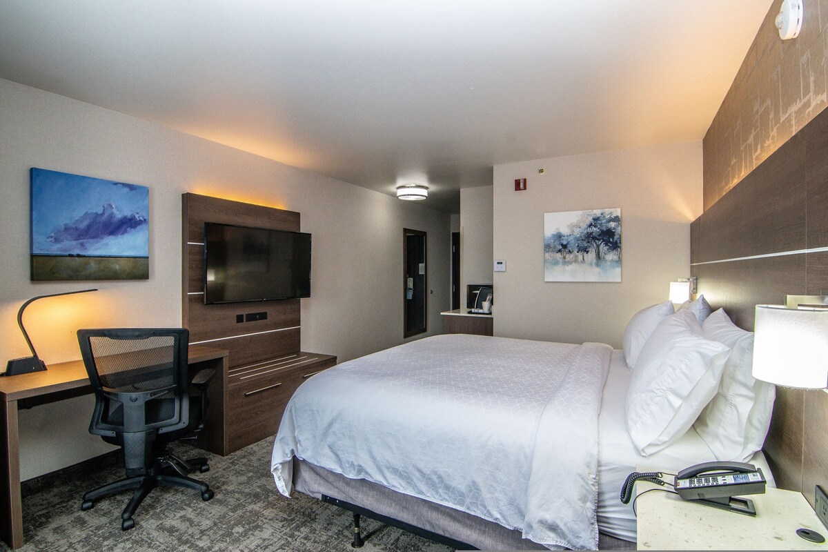 A hotel room with a bed, desk, chair, TV, and artwork on the walls.