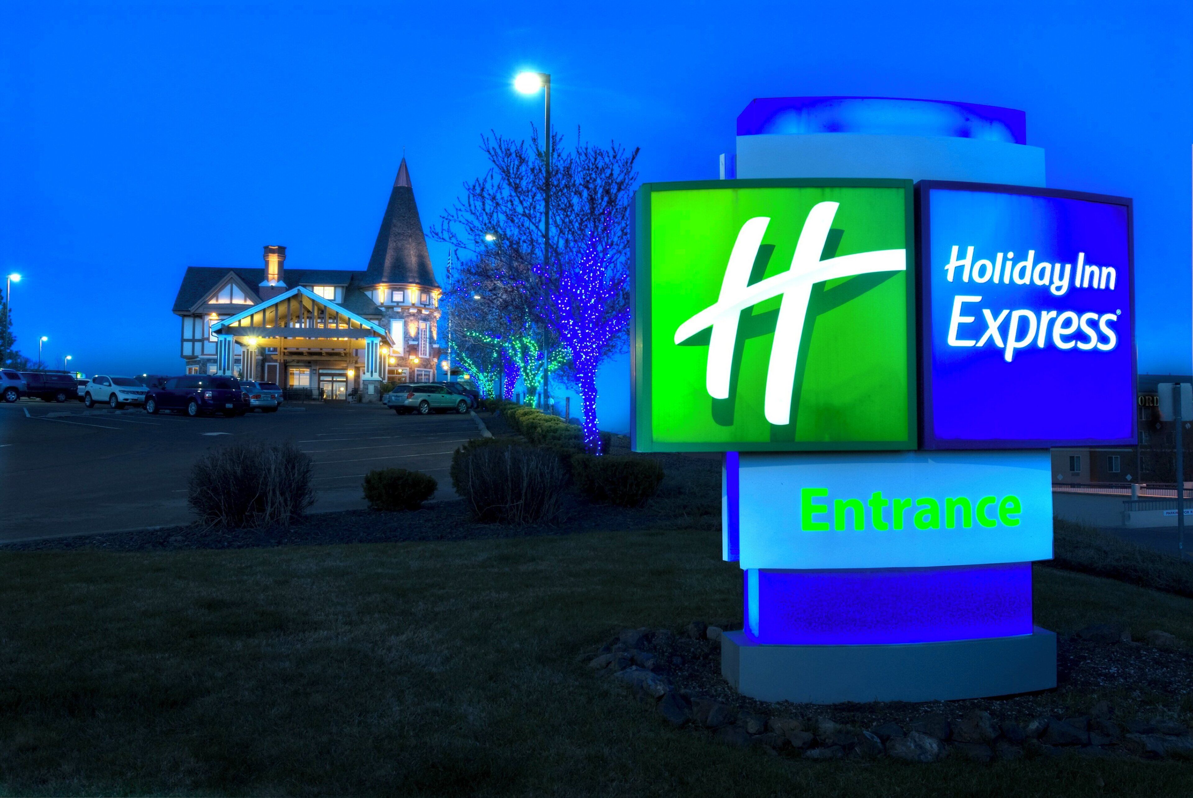 Ein Schild von Holiday Inn Express mit dem gut sichtbaren ‚H‘-Logo, einem Eingangsschild und einem Gebäude im Hintergrund.