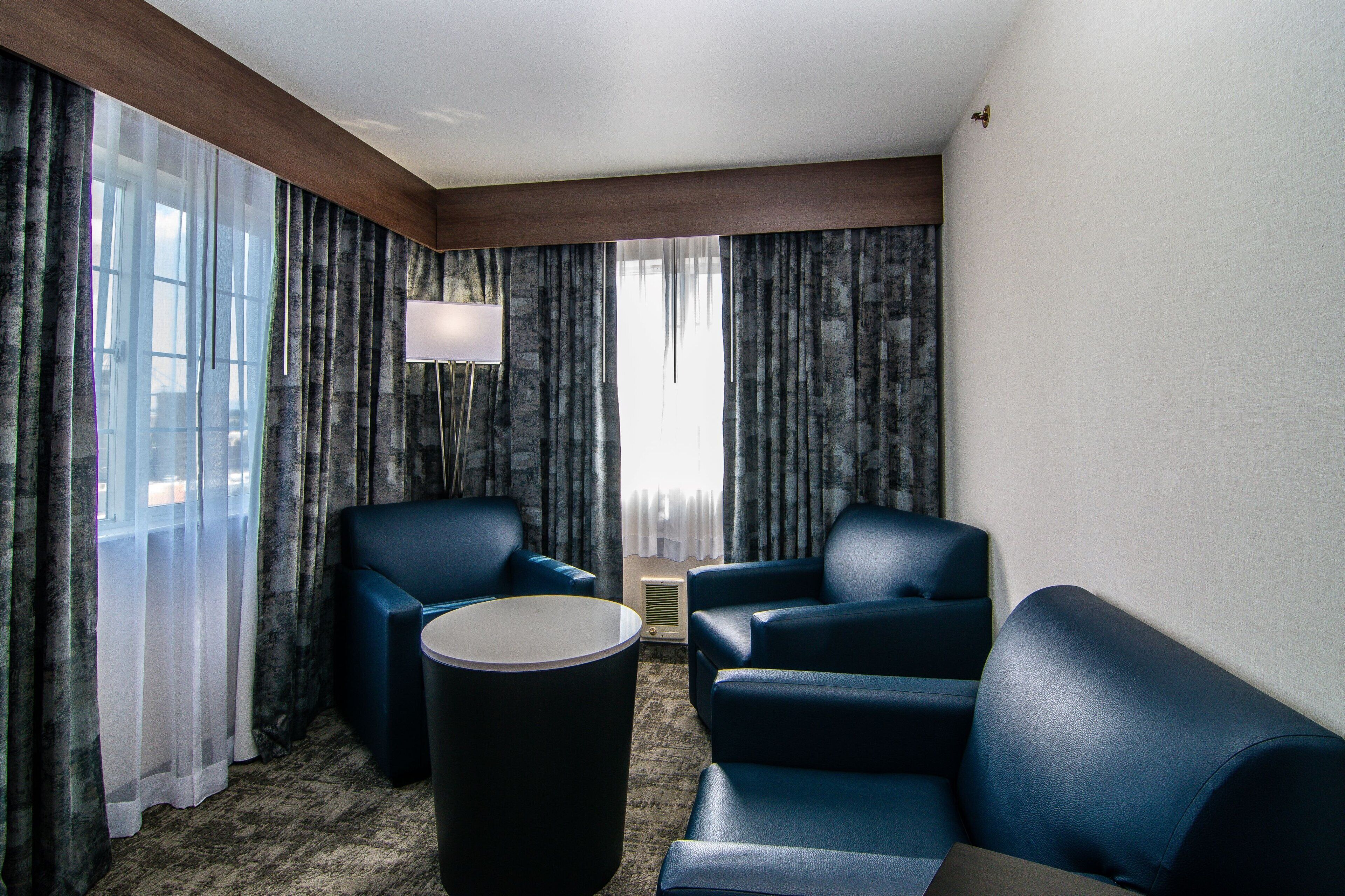 Quarto de hotel com duas poltronas azuis, uma mesa redonda pequena e uma janela grande com cortinas.