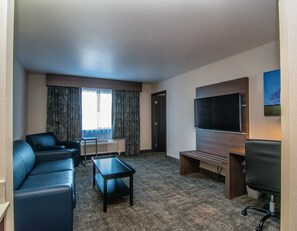 A hotel room with a seating area, a flat-screen TV, a window with curtains, and a carpeted floor.