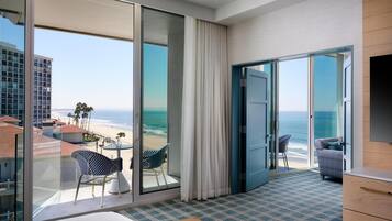 Habitación de hotel con un gran ventanal corredero que da a un balcón con vistas a la playa y al océano. Incluye una cama, una pequeña mesa con sillas y un televisor montado en la pared.