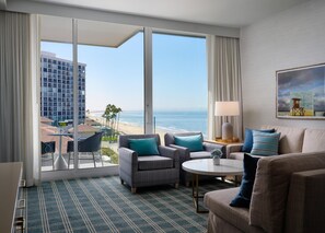 A hotel room with a large balcony, a sofa, a chair, a small table, and a painting on the wall.