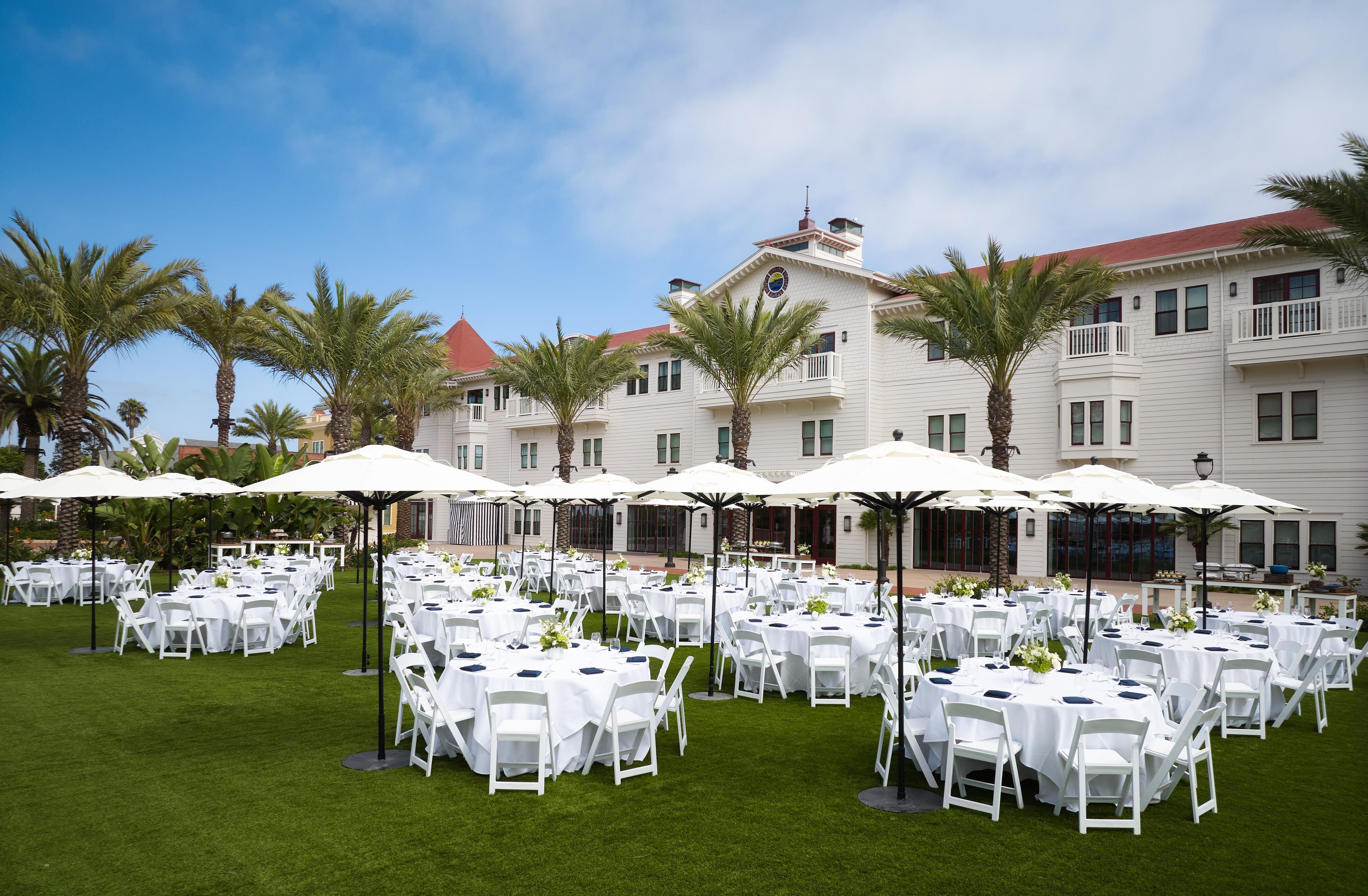 An outdoor event space with multiple round tables and chairs, surrounded by palm trees and umbrellas.