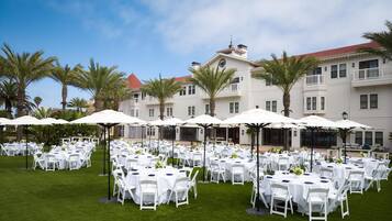 An outdoor event space with multiple round tables and chairs, surrounded by palm trees and umbrellas.
