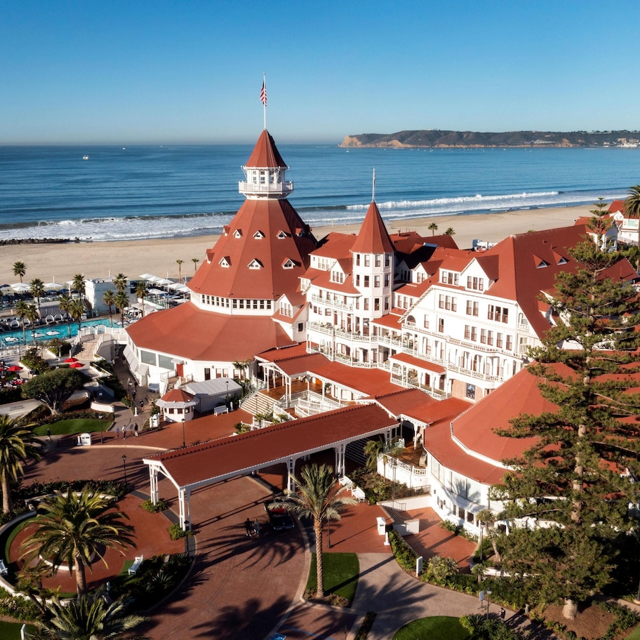 A large hotel with multiple red-roofed buildings, a swimming pool, and a beach view.