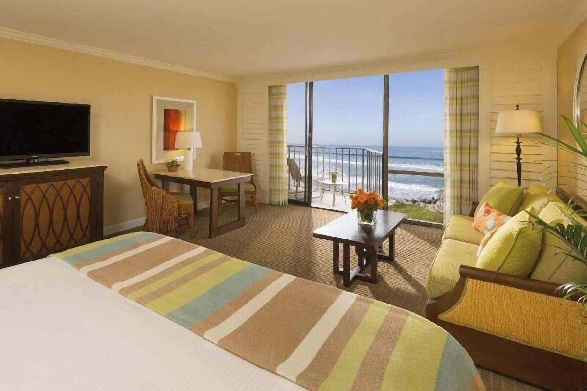 A hotel room with a bed, a television, a desk, a chair, a sofa, a table with flowers, and a balcony with a view of the ocean.