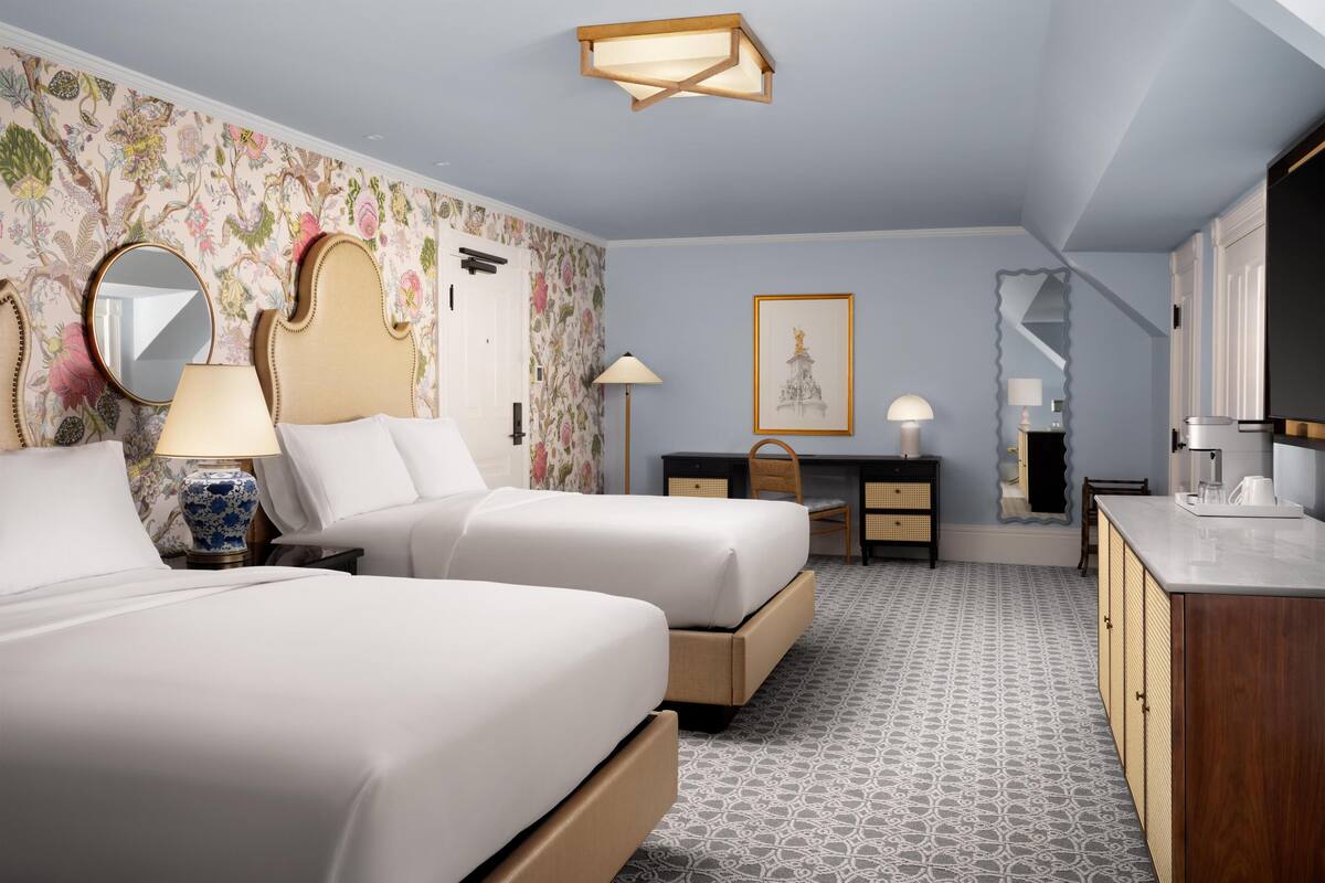 Une chambre d’hôtel avec deux lits, un bureau, une chaise, une armoire, un miroir, une lampe et un tableau encadré au mur.