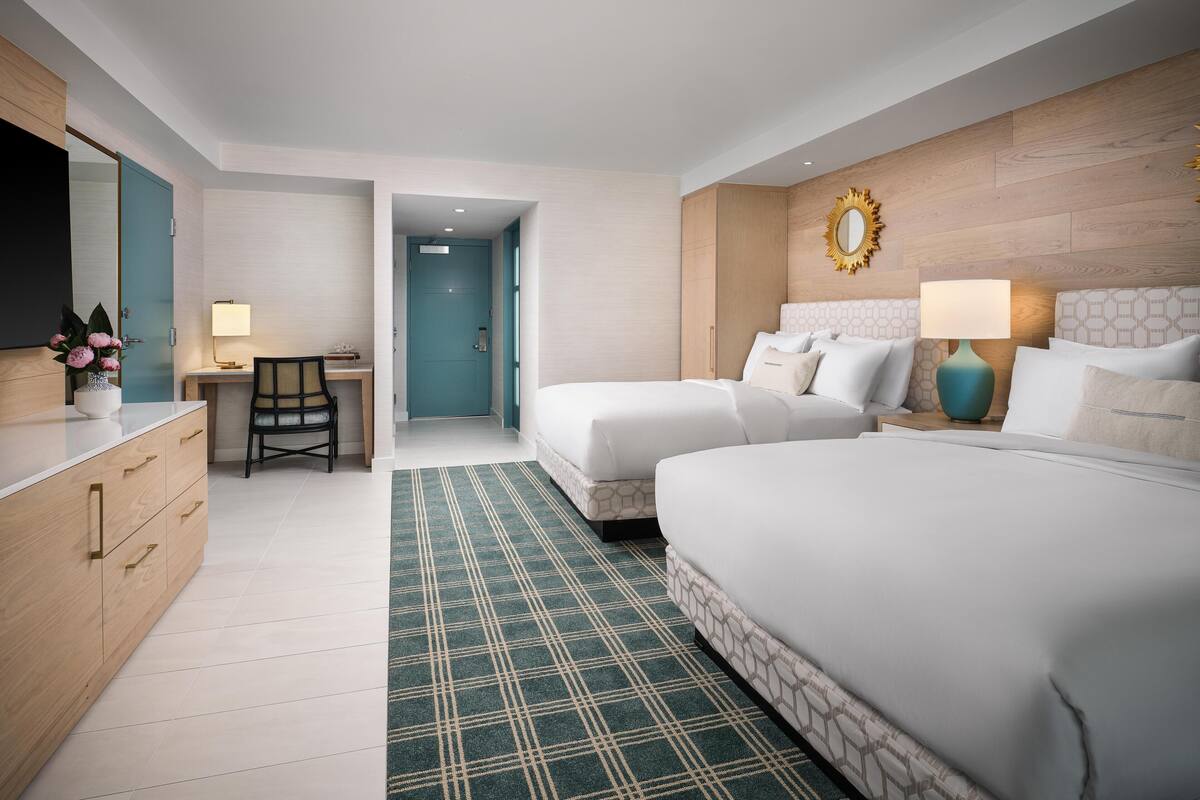 Une chambre d’hôtel équipée d’un bureau, de deux lits, d’un grand miroir et d’un tapis à motifs.