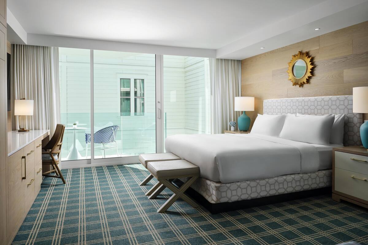 Une chambre d’hôtel avec un grand lit, deux tables de chevet avec des lampes, une chaise, une petite table avec une plante et une vue sur un immeuble par la fenêtre.