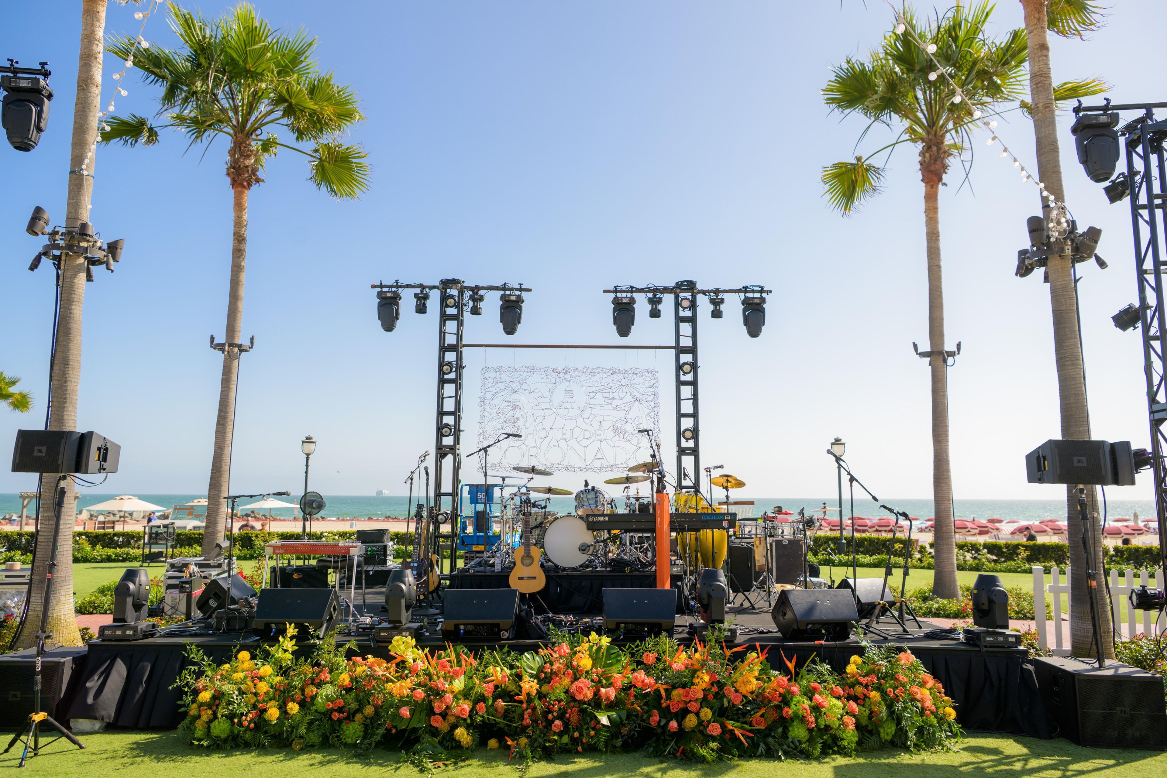 An outdoor stage with musical instruments and lighting equipment, surrounded by palm trees and a beach view.