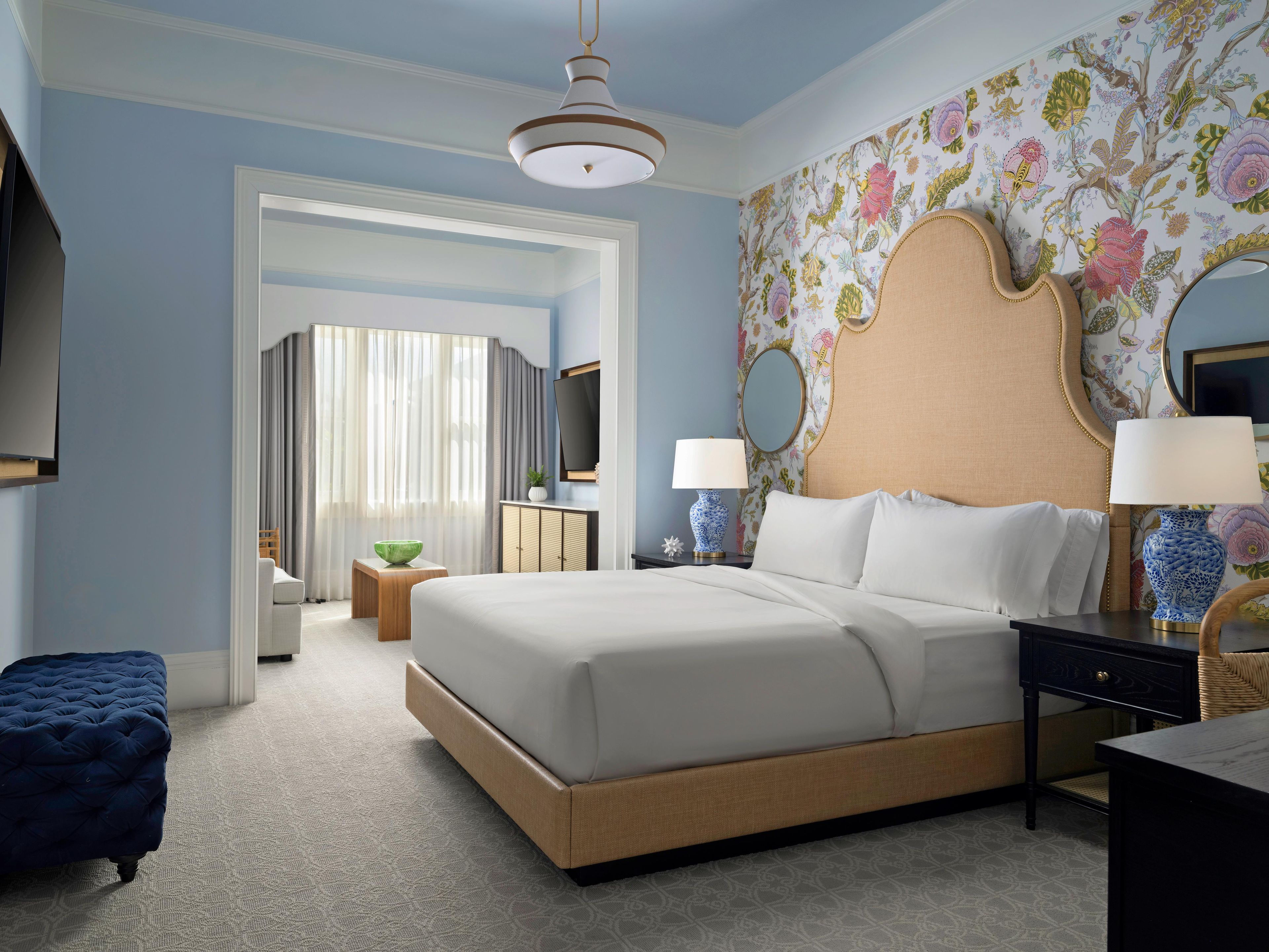 A well-appointed hotel room with a large bed, a nightstand, a dresser, a mirror, and a seating area with a blue ottoman.