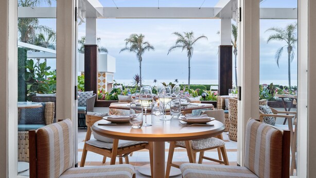 A dining area with a view of palm trees and the ocean, furnished with round tables and chairs. - Hotel del Coronado, Curio Collection by Hilton (Coronado)