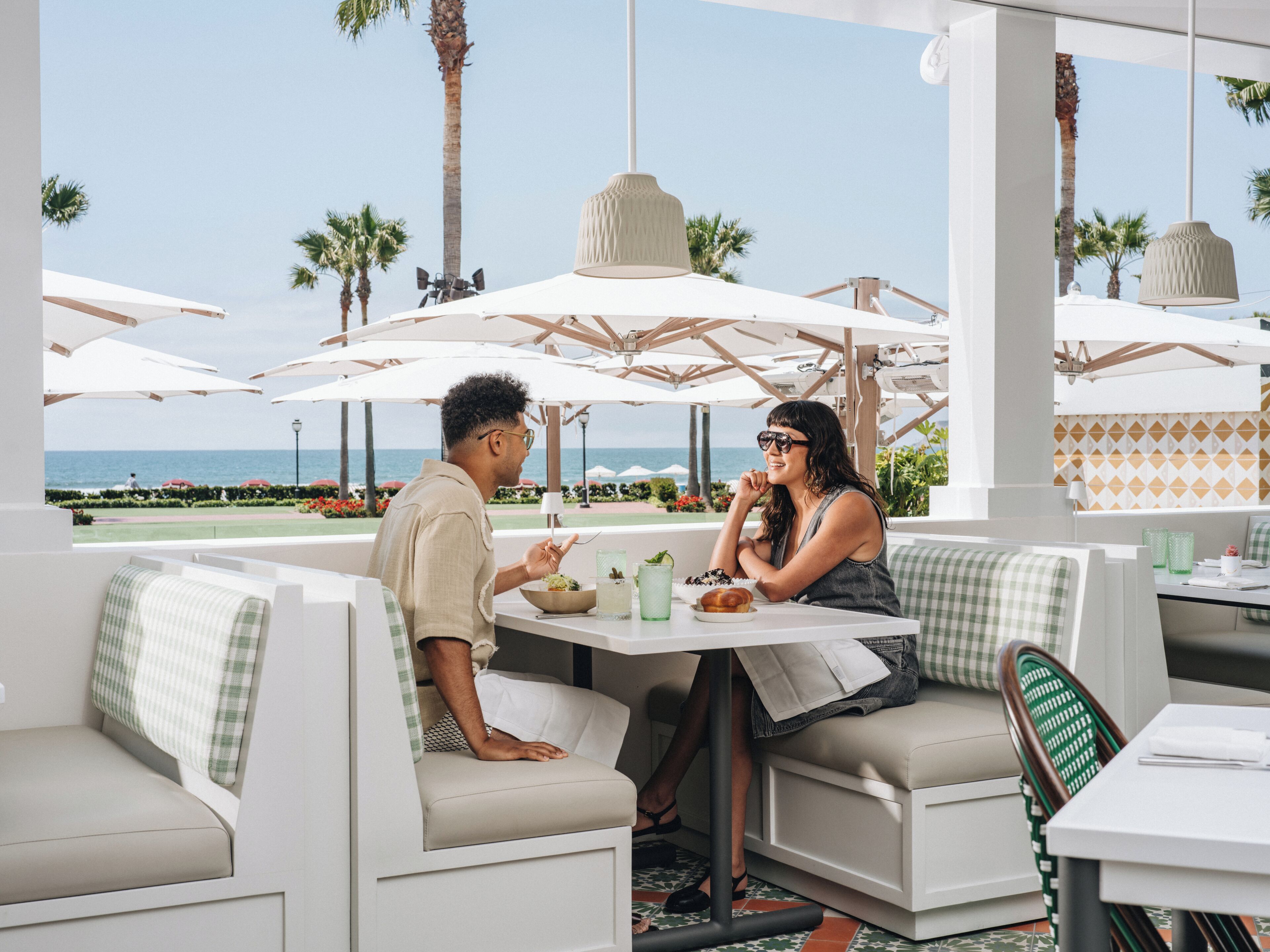 A couple dining at an outdoor cafe with a view of the ocean, white furniture, and umbrellas.