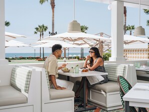 Ein Paar speist in einem Straßencafé mit Blick auf das Meer, weißes Mobiliar und Sonnenschirme.