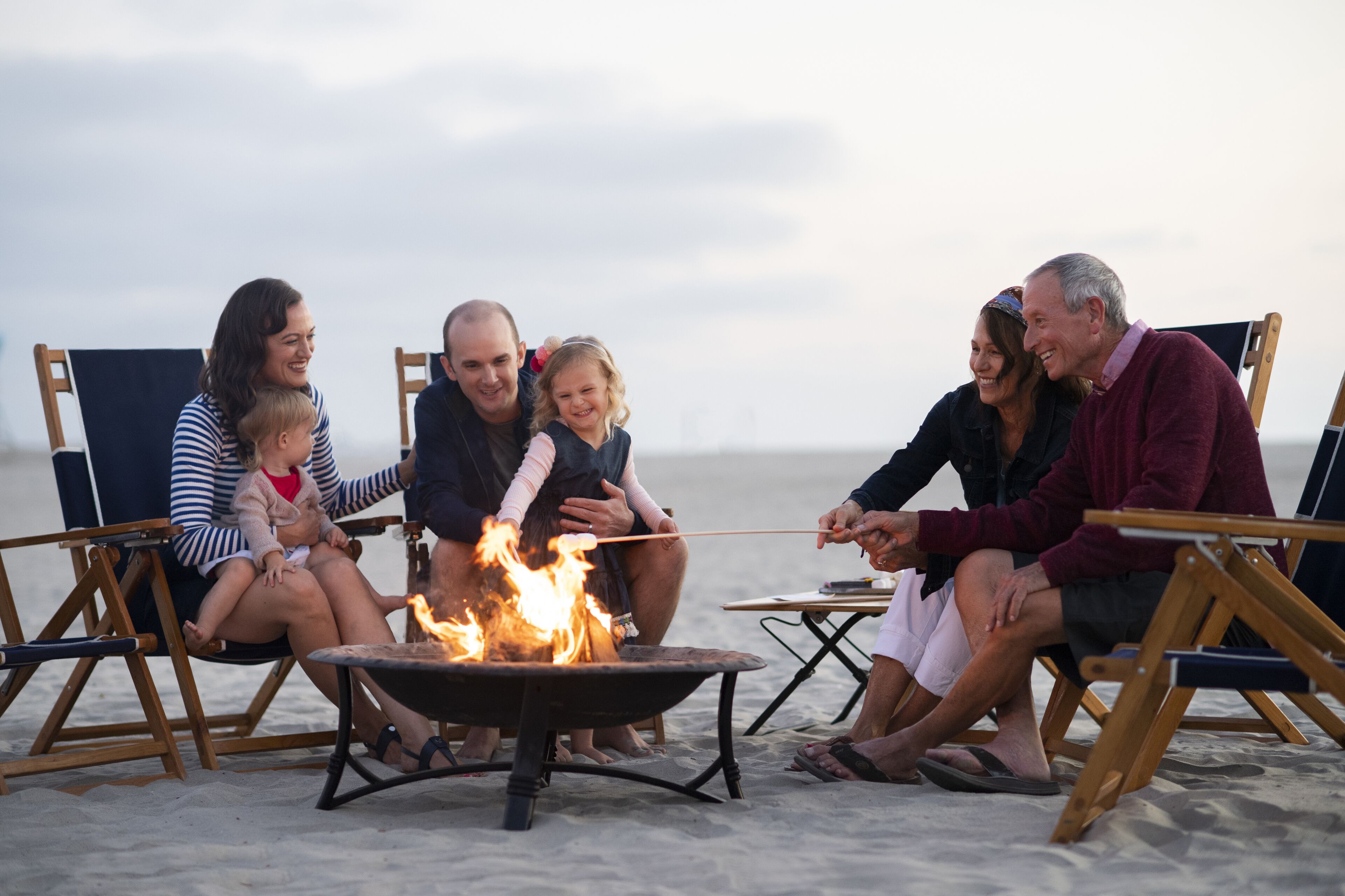 A group of people sitting on beach chairs around a campfire on a sandy beach.