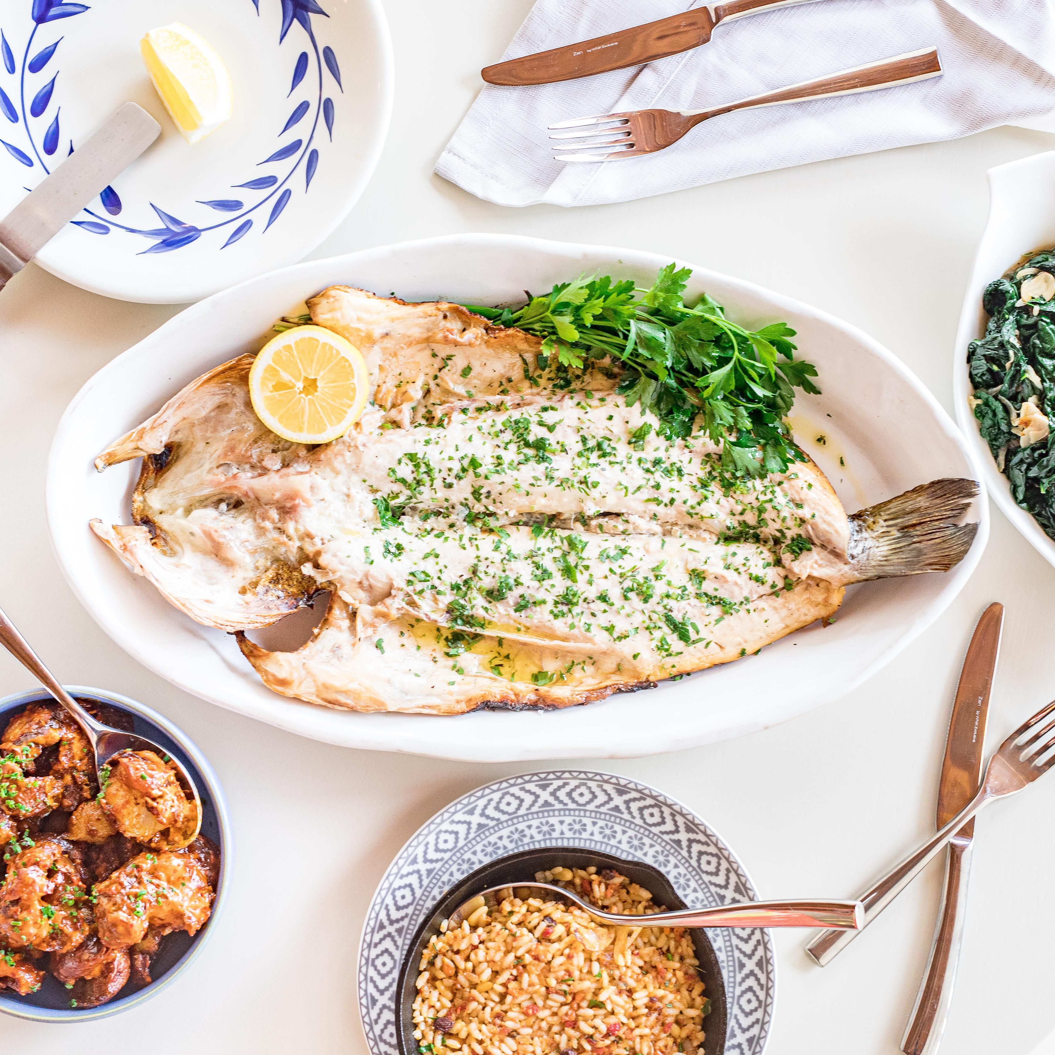 Prato branco com peixe, adornado com salsa e limão, acompanhado de uma tigela de arroz e um pequeno recipiente com molho.